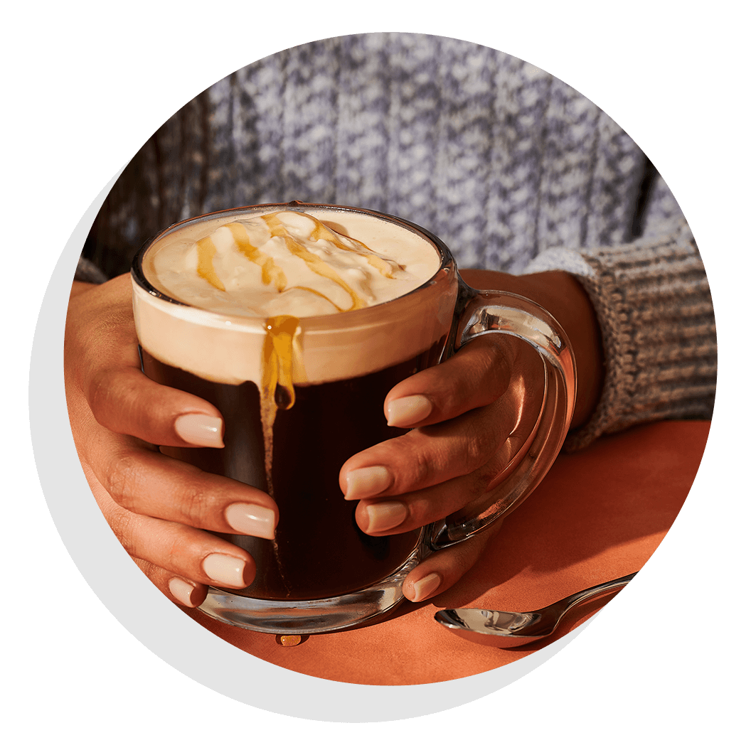 Hands holding clear mug of coffee with a foam top and honey drizzle.