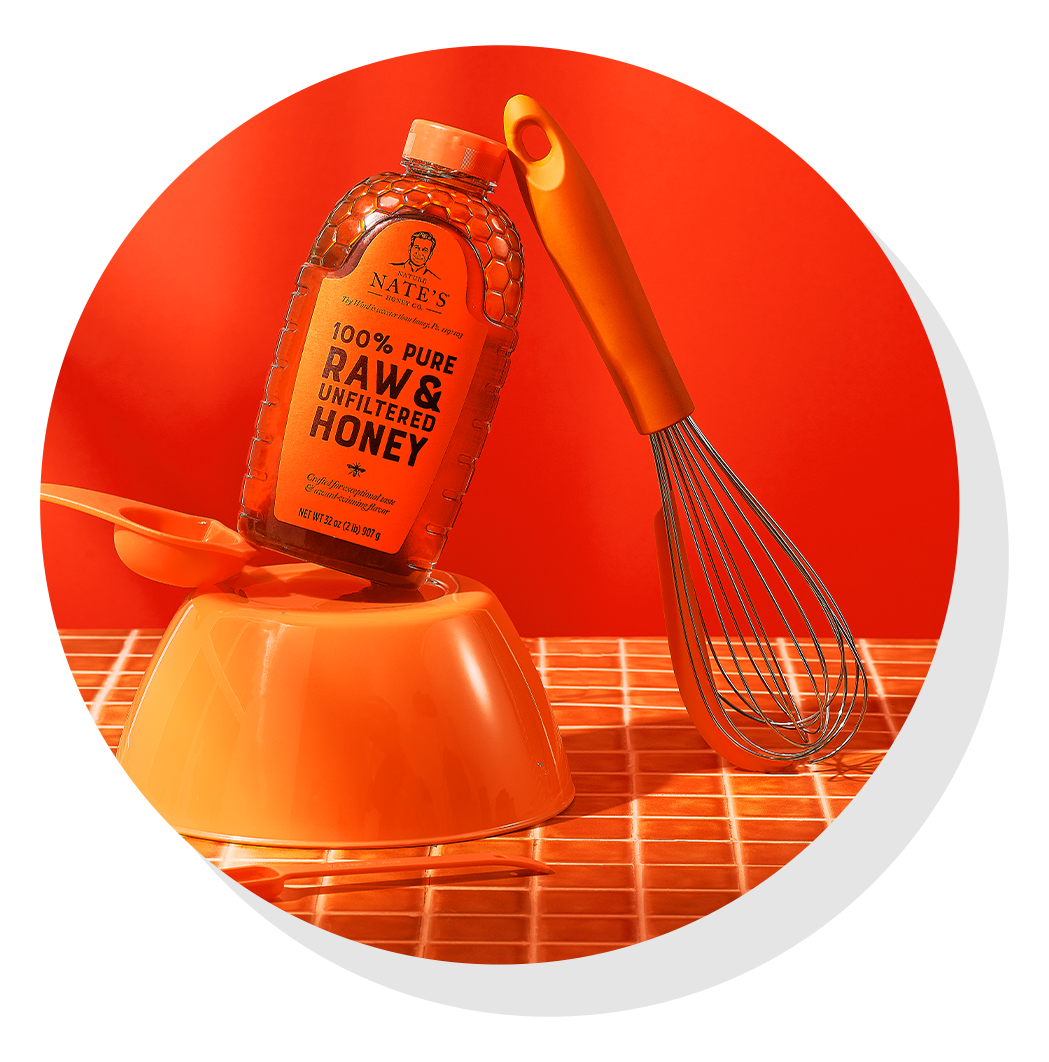 Nate’s honey bottle on top of an orange mixing bowl beside a whisk, set against an orange background.
