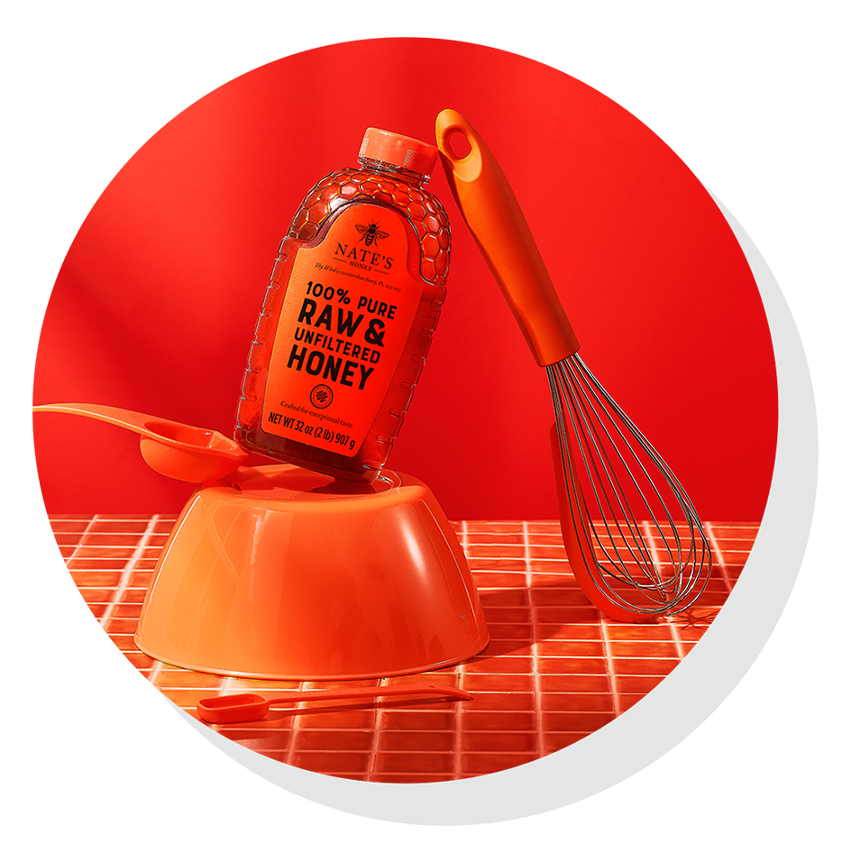 Nate’s honey bottle on top of an orange mixing bowl beside a whisk, set against an orange background.