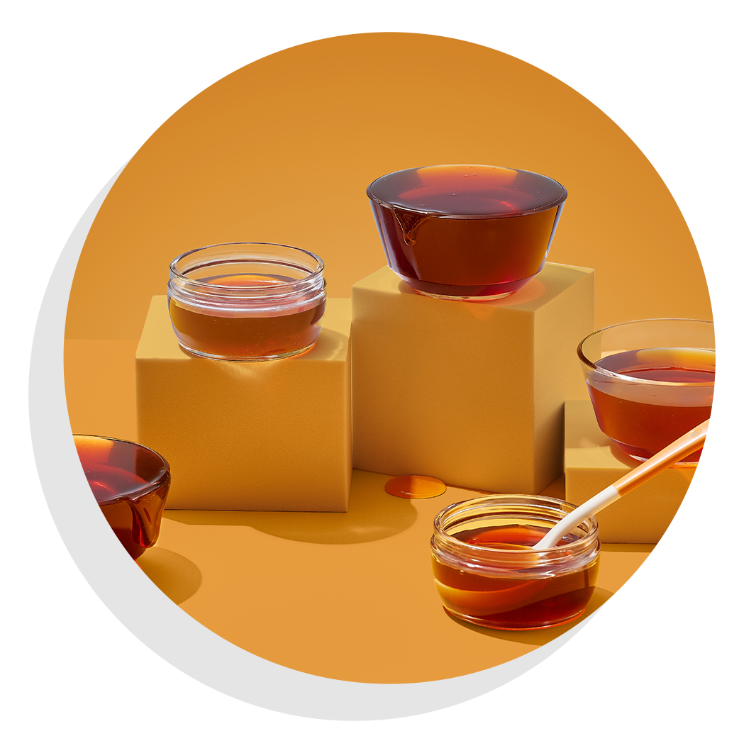 Small glass bowls of honey arranged on stacked blocks against a warm golden background, with a honey dipper resting in one bowl.