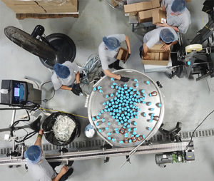 Top view of workers in a factory sorting blue capsules on a large metal table, with equipment and boxes around.